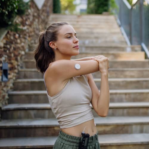 A woman in workout clothes stretches her arm outdoors in FL. She wears a glucose monitor on her upper arm and an insulin pump clipped to her waistband, standing on stairs with greenery in the background—highlighting active primary care Tallahassee living.