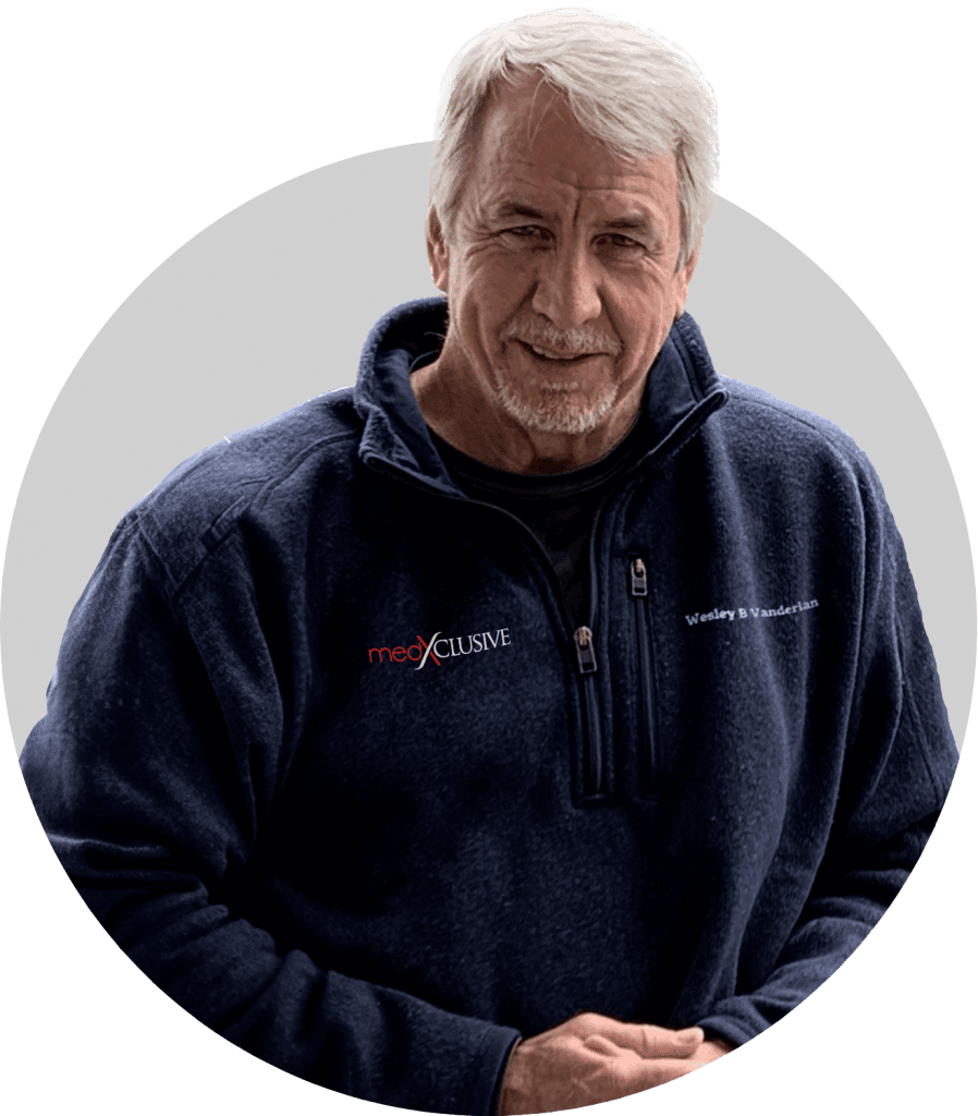 An older man with gray hair and a short beard wears a navy blue zip-up sweater with "medXCLUSIVE" and "Wesley B Vandeaarden" embroidered on it, representing primary care in Tallahassee, FL, as he stands smiling gently against a light background.