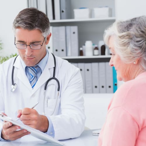 A male doctor in a white coat writes on a clipboard while talking to an older woman with gray hair in a pink sweater. They sit in a bright primary care Tallahassee, FL office, with shelves and files visible in the background.