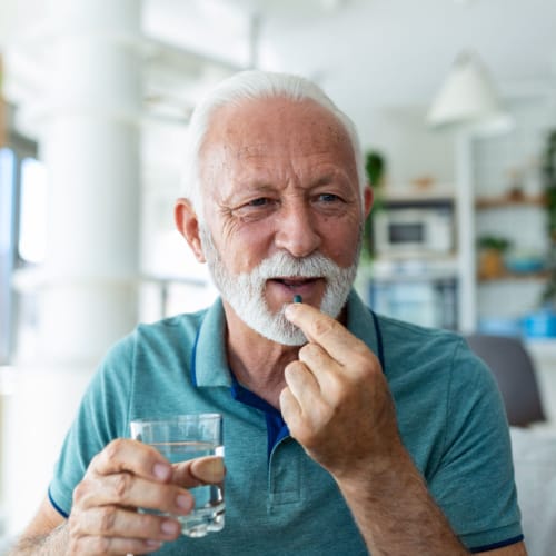 An older man with a white beard holds a glass of water in one hand and is about to take a pill with the other, smiling in his bright, modern FL home—showcasing the benefits of quality primary care Tallahassee residents trust.