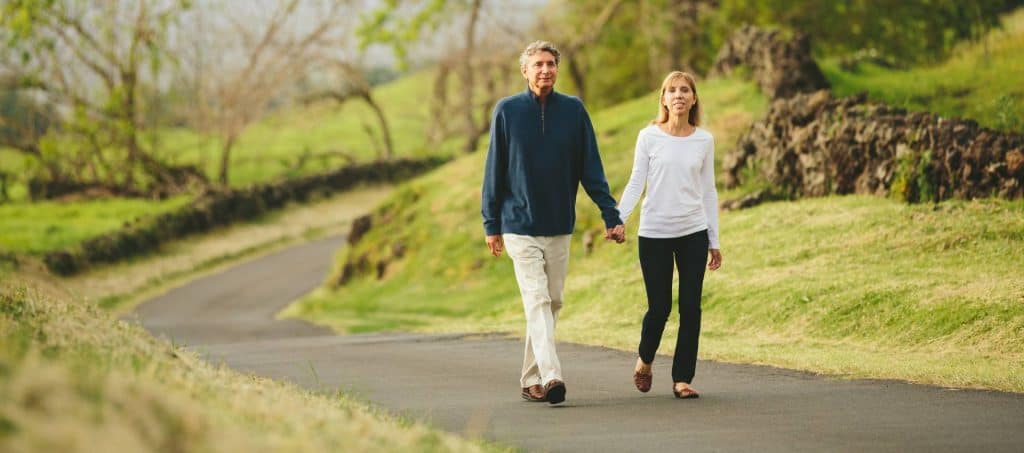 An older couple is walking hand in hand on a paved path through a green, scenic FL park with trees and grass on either side.
