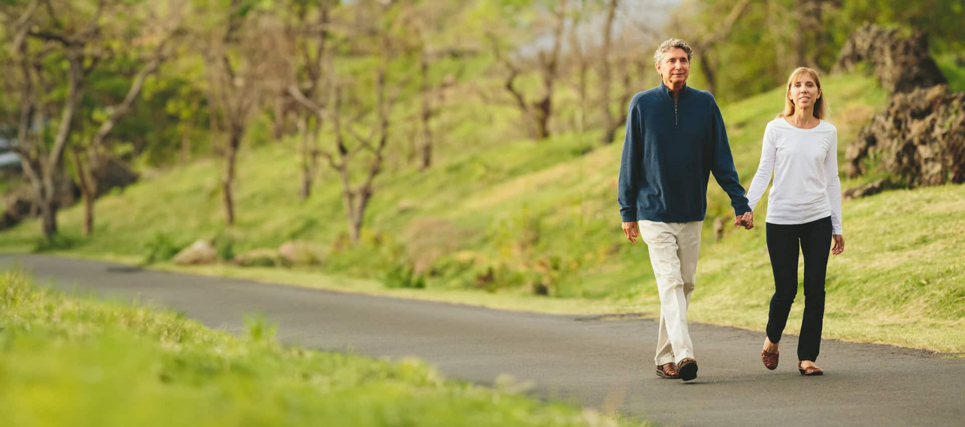 A man and woman walk hand in hand along a paved path in a Tallahassee park, surrounded by green grass and trees on a sunny FL day.