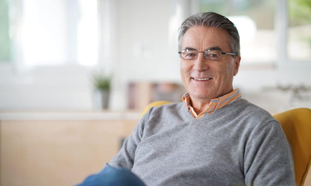 A middle-aged man with gray hair and glasses, wearing a gray sweater over an orange collared shirt, smiles while sitting on a yellow chair in a bright, modern primary care Tallahassee, FL office.
