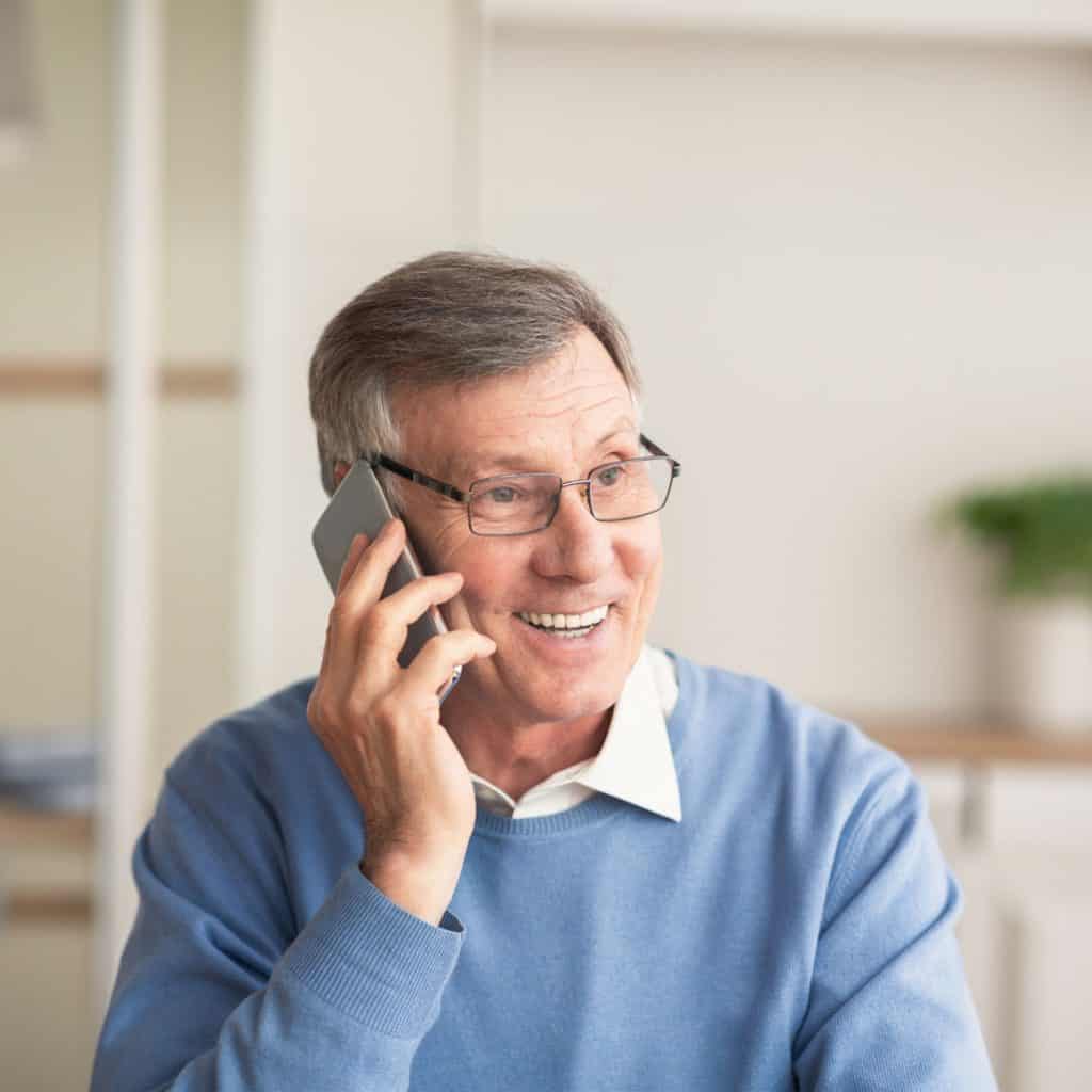 Smiling older man with glasses, wearing a blue sweater and white shirt, talks on a smartphone indoors—perhaps scheduling an appointment for primary care in Tallahassee, FL.