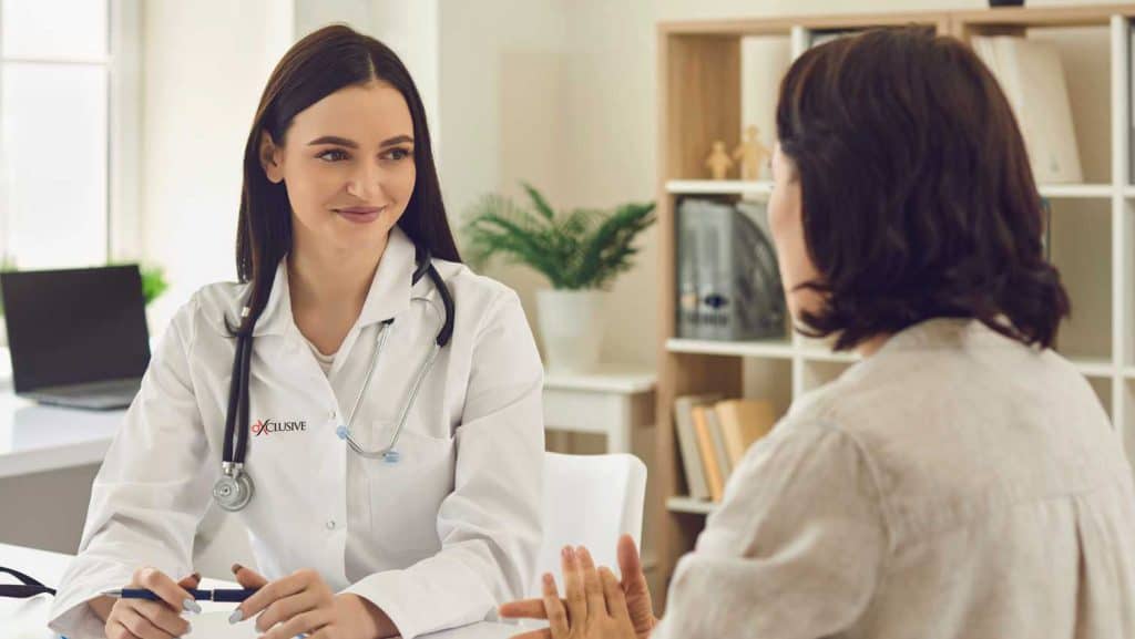 A female doctor in a white coat with a stethoscope around her neck smiles while talking to a patient across a desk in a bright, modern FL primary care Tallahassee medical office.