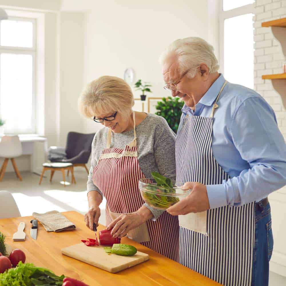 An elderly couple wearing aprons prepares vegetables together in a bright FL kitchen; the woman slices a red bell pepper while the man, smiling, holds a bowl of leafy greens—enjoying healthy habits supported by primary care Tallahassee.