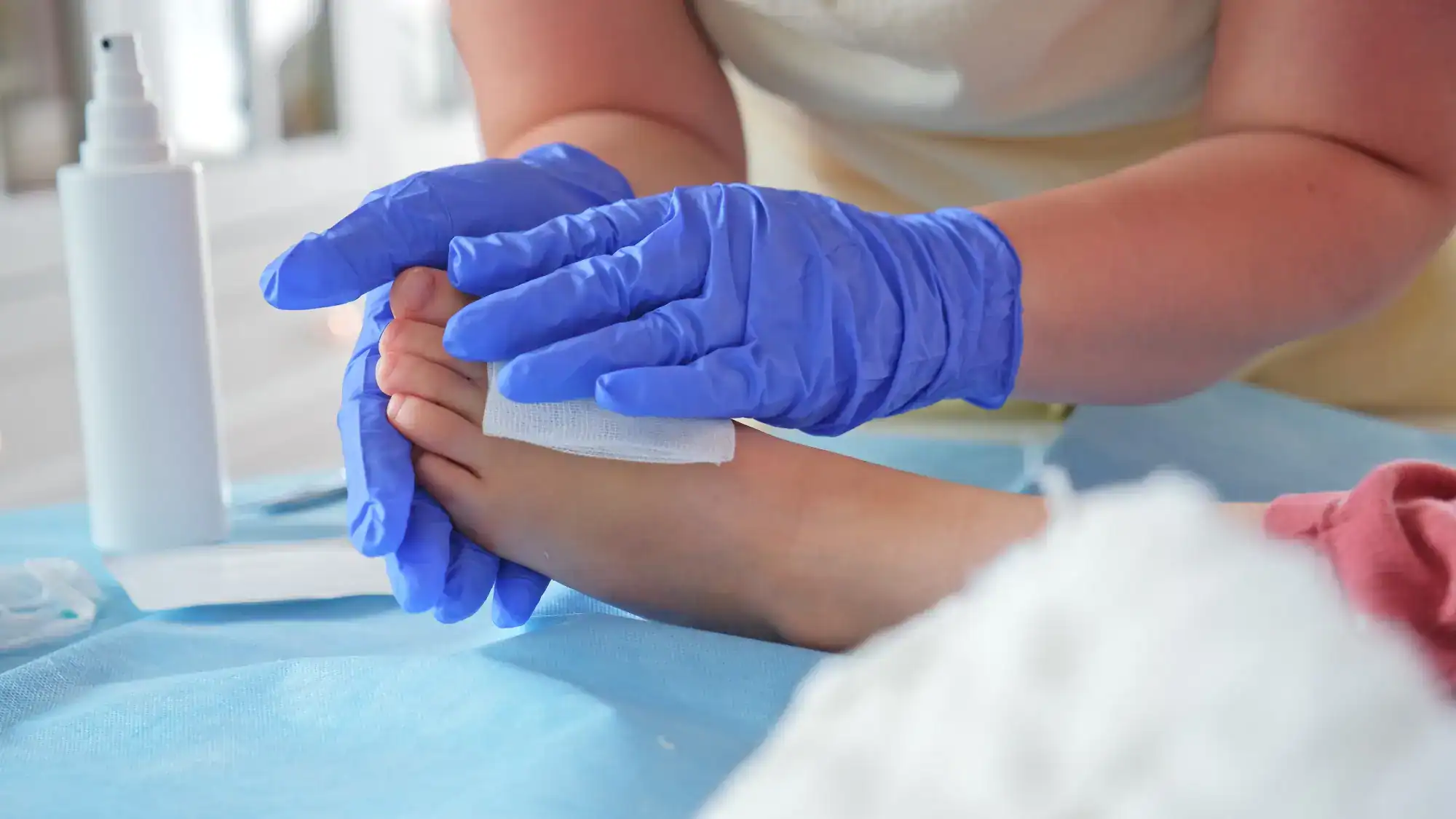 A person wearing blue gloves cleans another person's foot with a white gauze pad, while a spray bottle rests nearby on a blue surface—capturing attentive care, often seen in primary care Tallahassee clinics in FL.