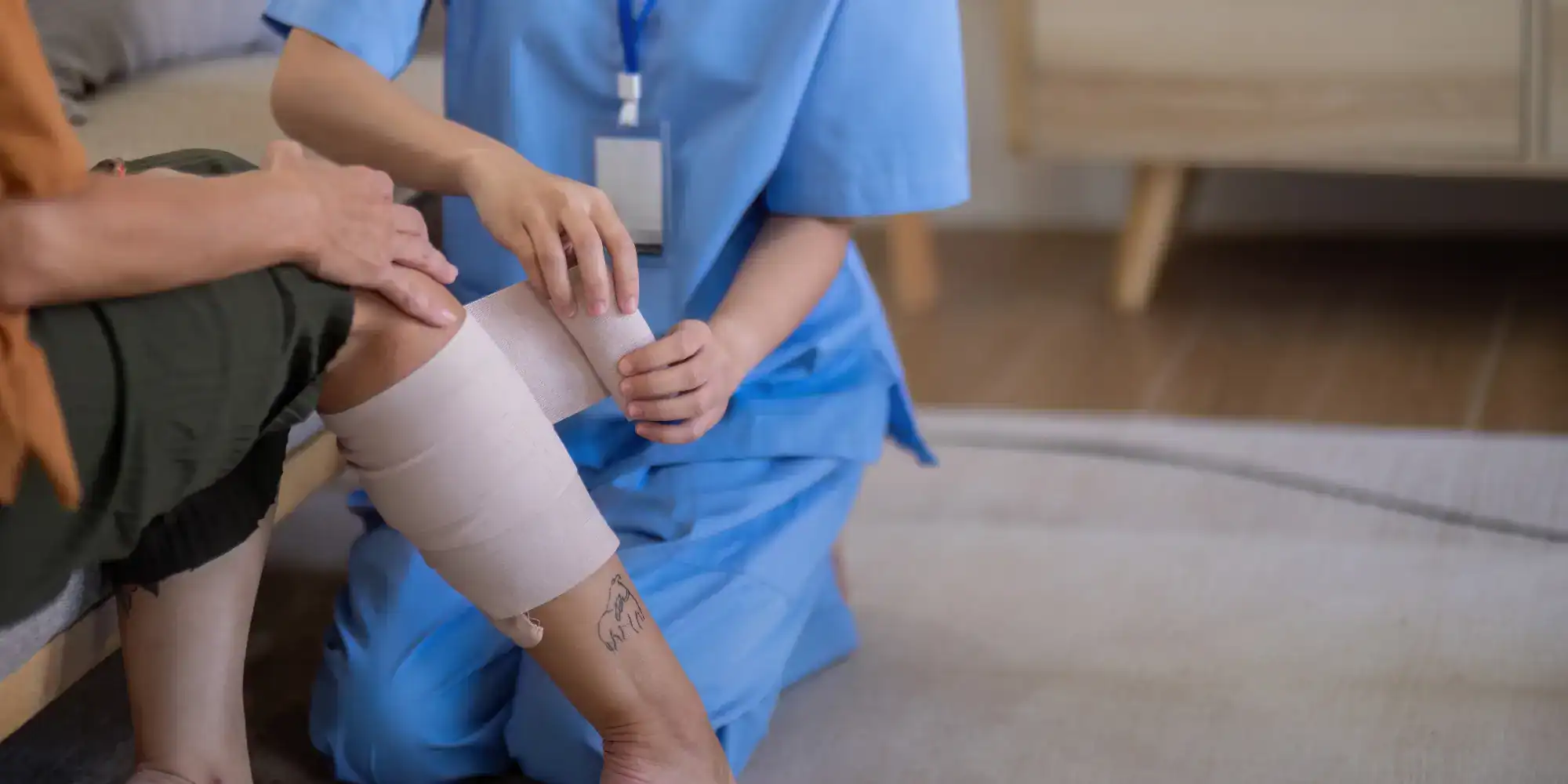A healthcare worker in blue scrubs wraps a bandage around a patient's tattooed, partially bent leg while sitting on the floor indoors, providing primary care Tallahassee patients can trust.