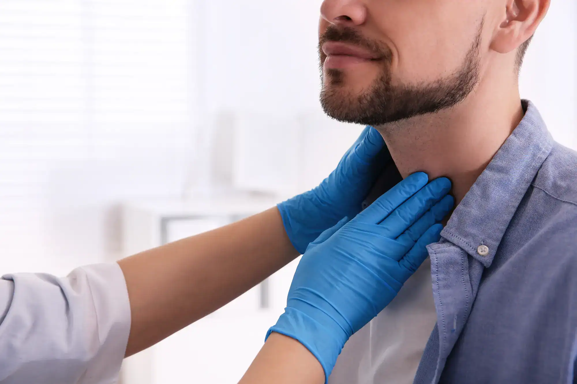 A primary care provider in Tallahassee, FL, wearing blue gloves, examines a man's throat by gently pressing on his neck—likely checking his thyroid gland. The bearded man is dressed in a blue shirt.