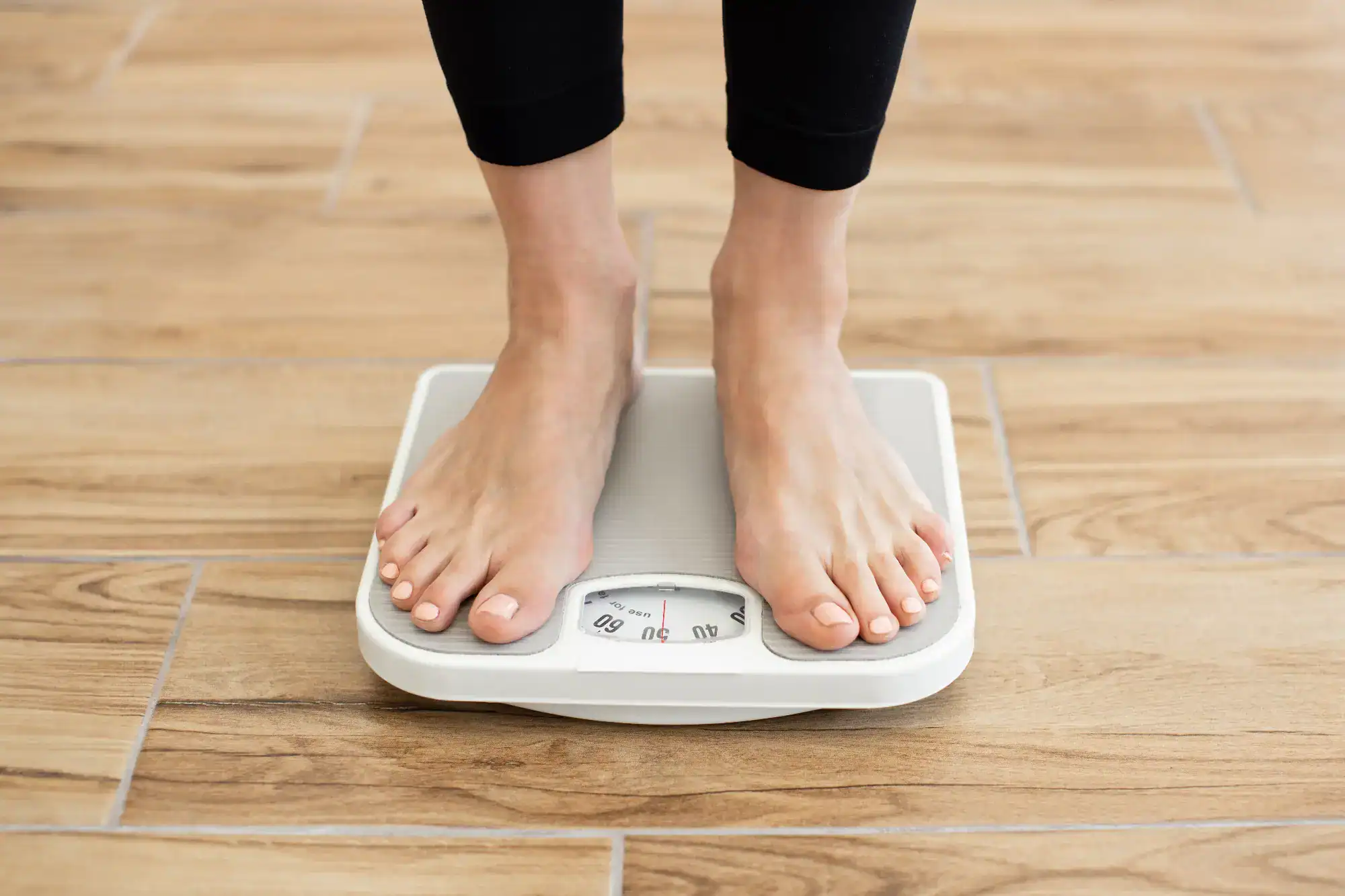 A person standing barefoot on a white analog bathroom scale placed on a wooden floor in FL, with the scale's dial visible between their feet—an image often seen in primary care Tallahassee clinics.
