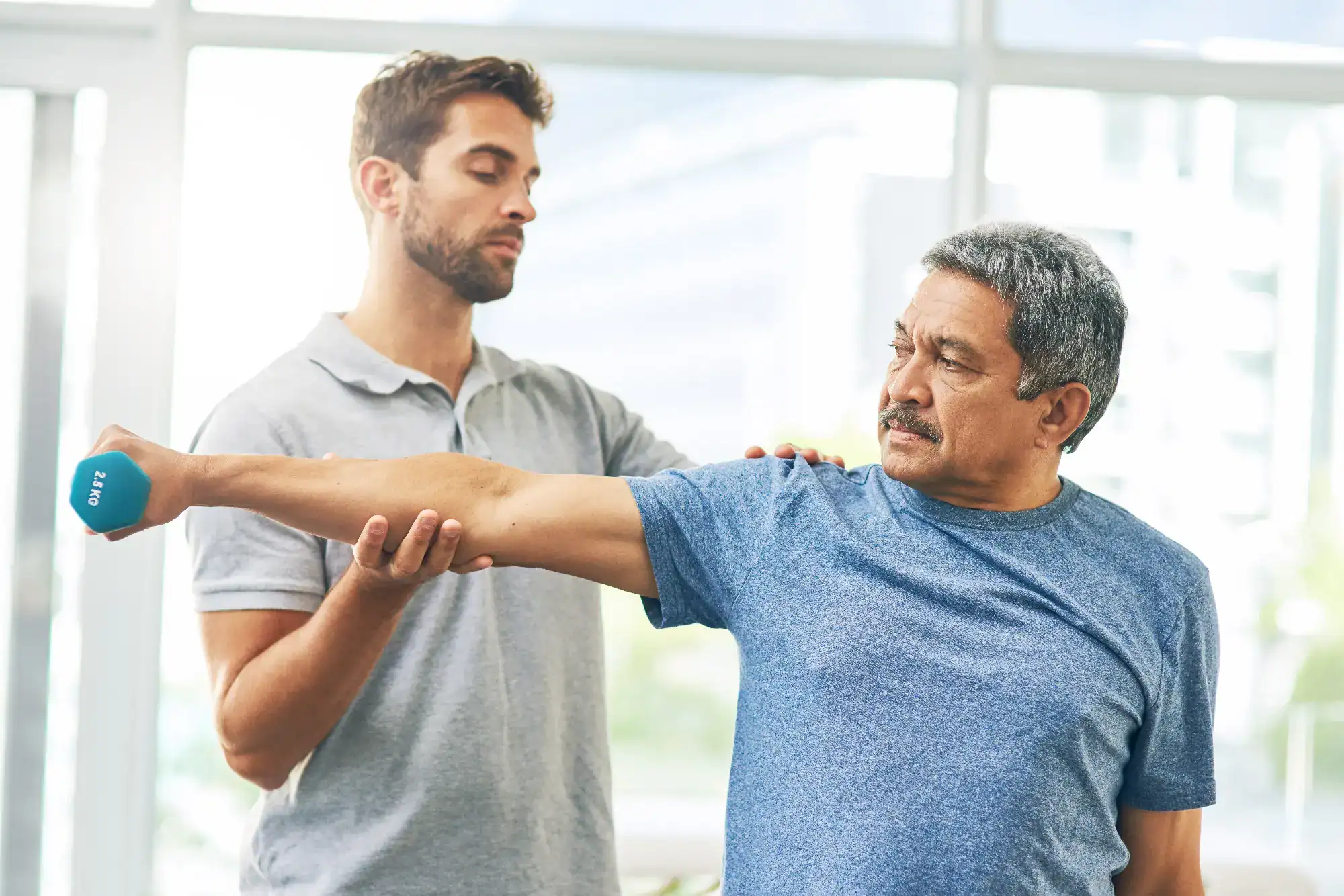 A man in a blue shirt lifts a dumbbell out to his side while another man assists at the shoulder, likely during a physical therapy session at a FL primary care Tallahassee clinic.