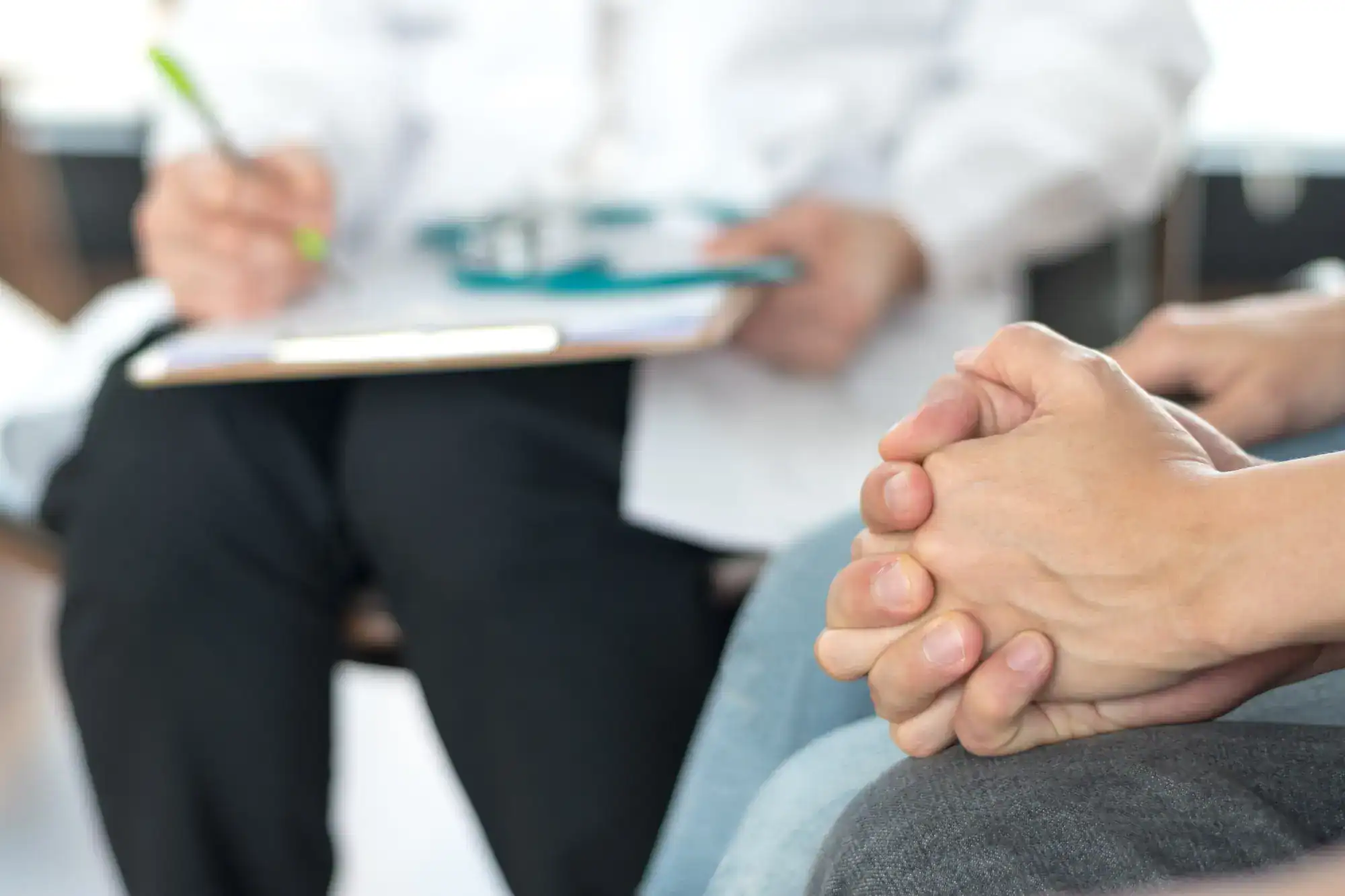 Close-up of a person's clasped hands resting on their lap, with a primary care Tallahassee doctor in a white coat holding a clipboard and pen in the background, suggesting a medical consultation or discussion in FL.