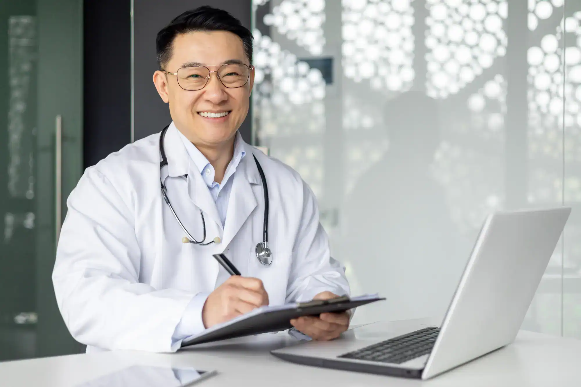 A smiling doctor in a white coat with a stethoscope around his neck writes on a clipboard while sitting at a desk with a laptop in a bright, modern primary care Tallahassee, FL office.