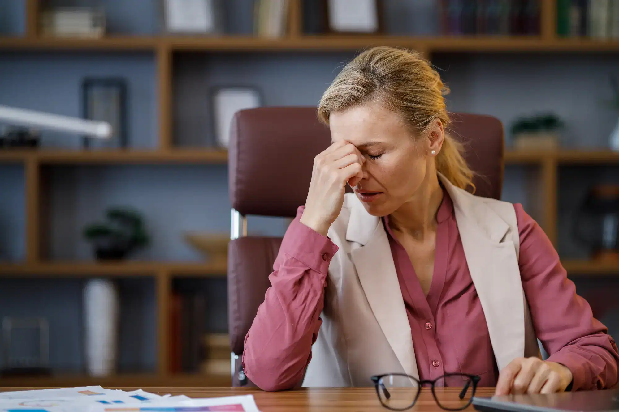A woman in business attire sits at a desk, pinching the bridge of her nose with her eyes closed, appearing stressed or tired—a scene familiar in FL, especially among busy primary care Tallahassee professionals. Office shelves with books and decor are visible in the background.
