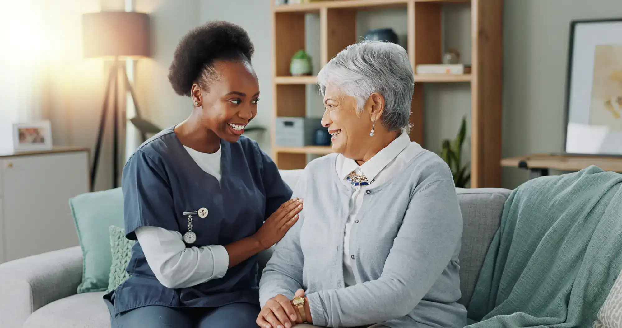 A nurse in scrubs smiles and gently holds the arm of an older woman with gray hair sitting on a couch, both looking at each other in a warmly lit living room, reflecting compassionate FL primary care in Tallahassee.