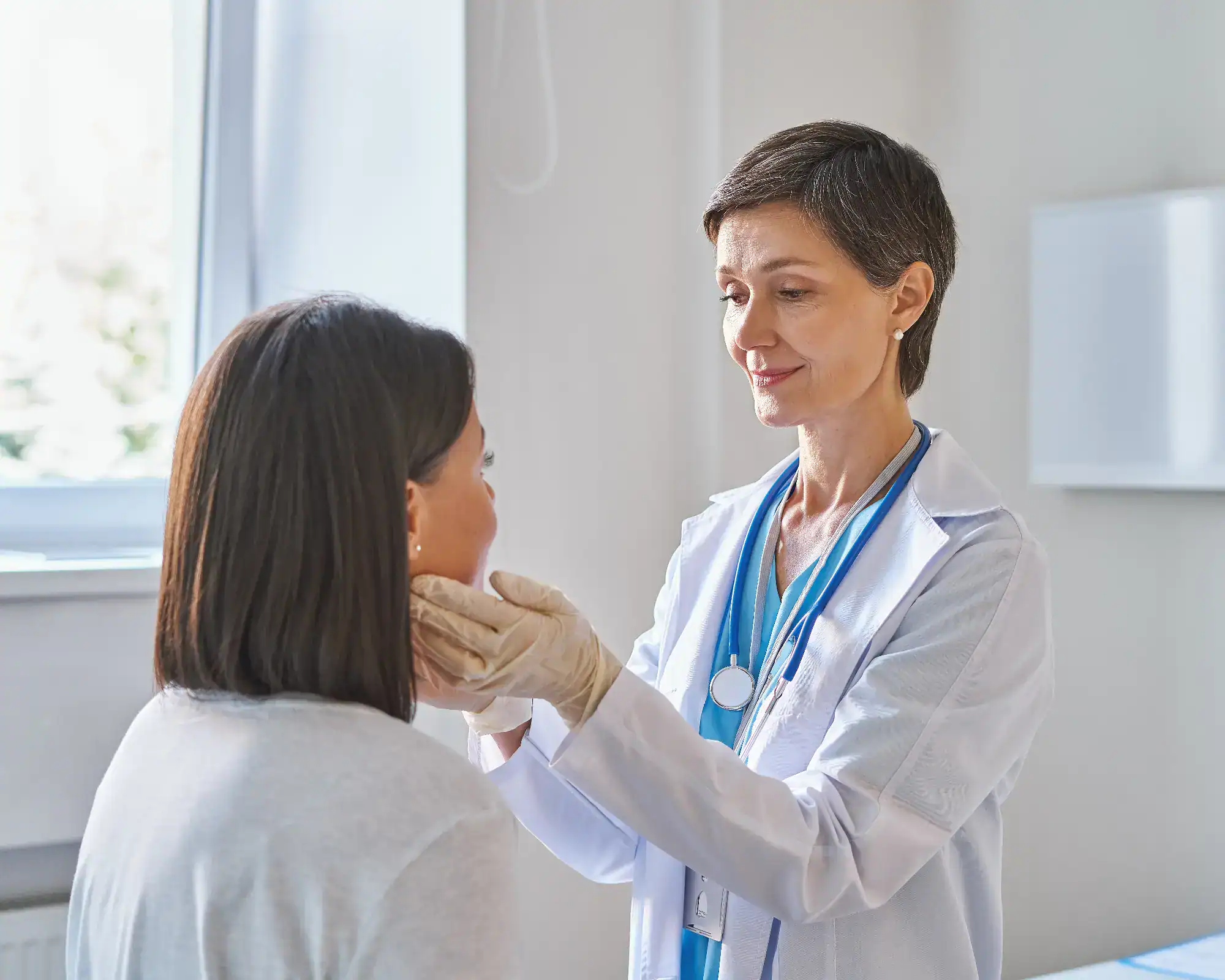 A doctor wearing a white coat and gloves examines a patient's neck in a bright primary care Tallahassee office. The patient is seated with her back to the camera.