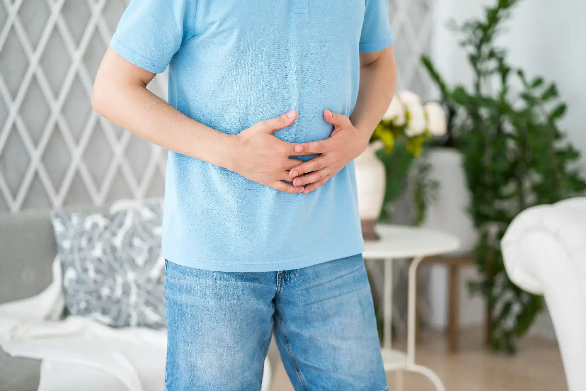 A person wearing a light blue shirt and jeans is holding their stomach with both hands, appearing to have abdominal discomfort, possibly needing primary care in Tallahassee, FL, while standing in a living room with plants and a table in the background.