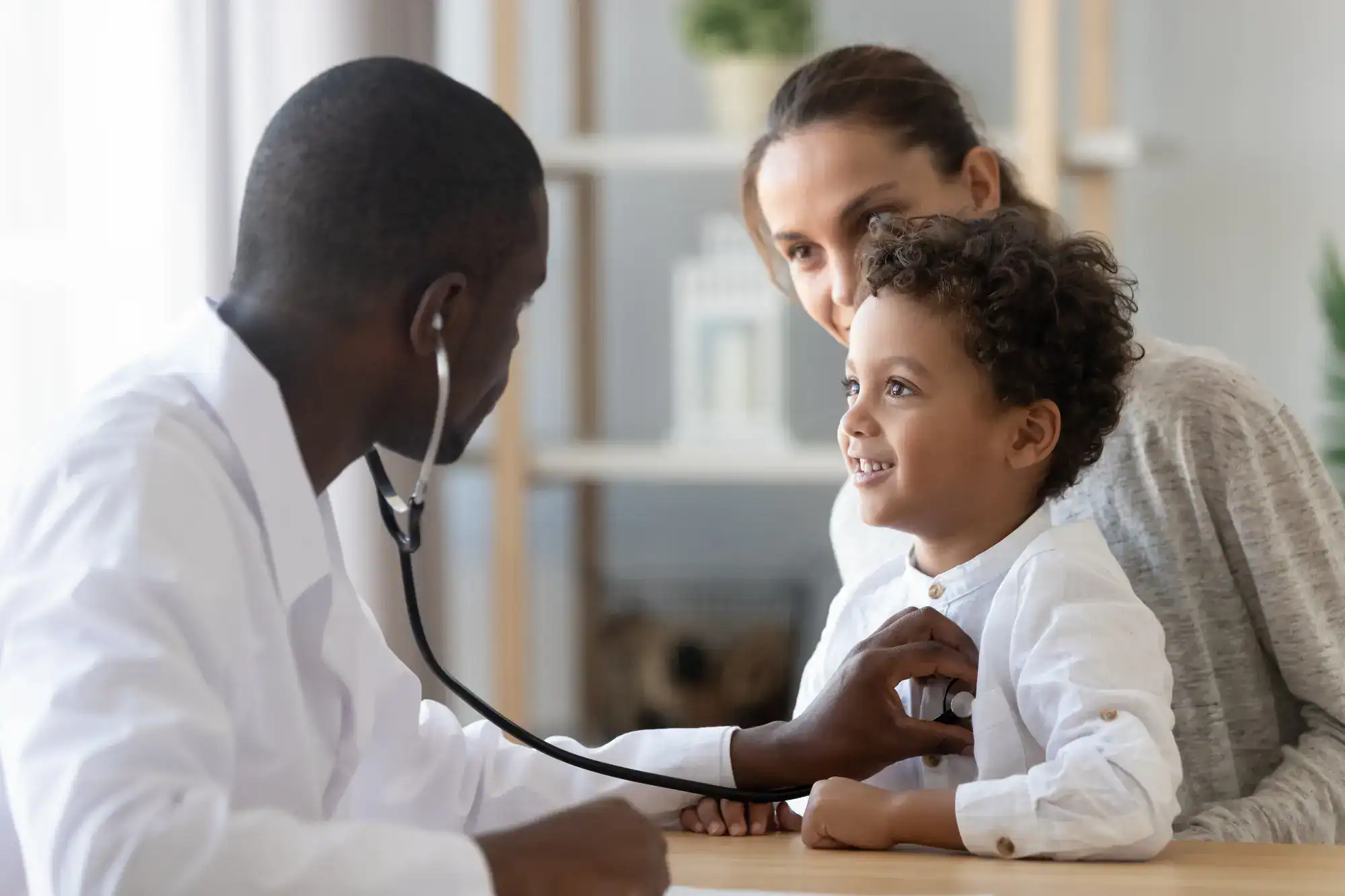 A doctor in FL uses a stethoscope to listen to a young boy's chest while the boy smiles and his mother looks on supportively in a bright, comfortable primary care Tallahassee office.