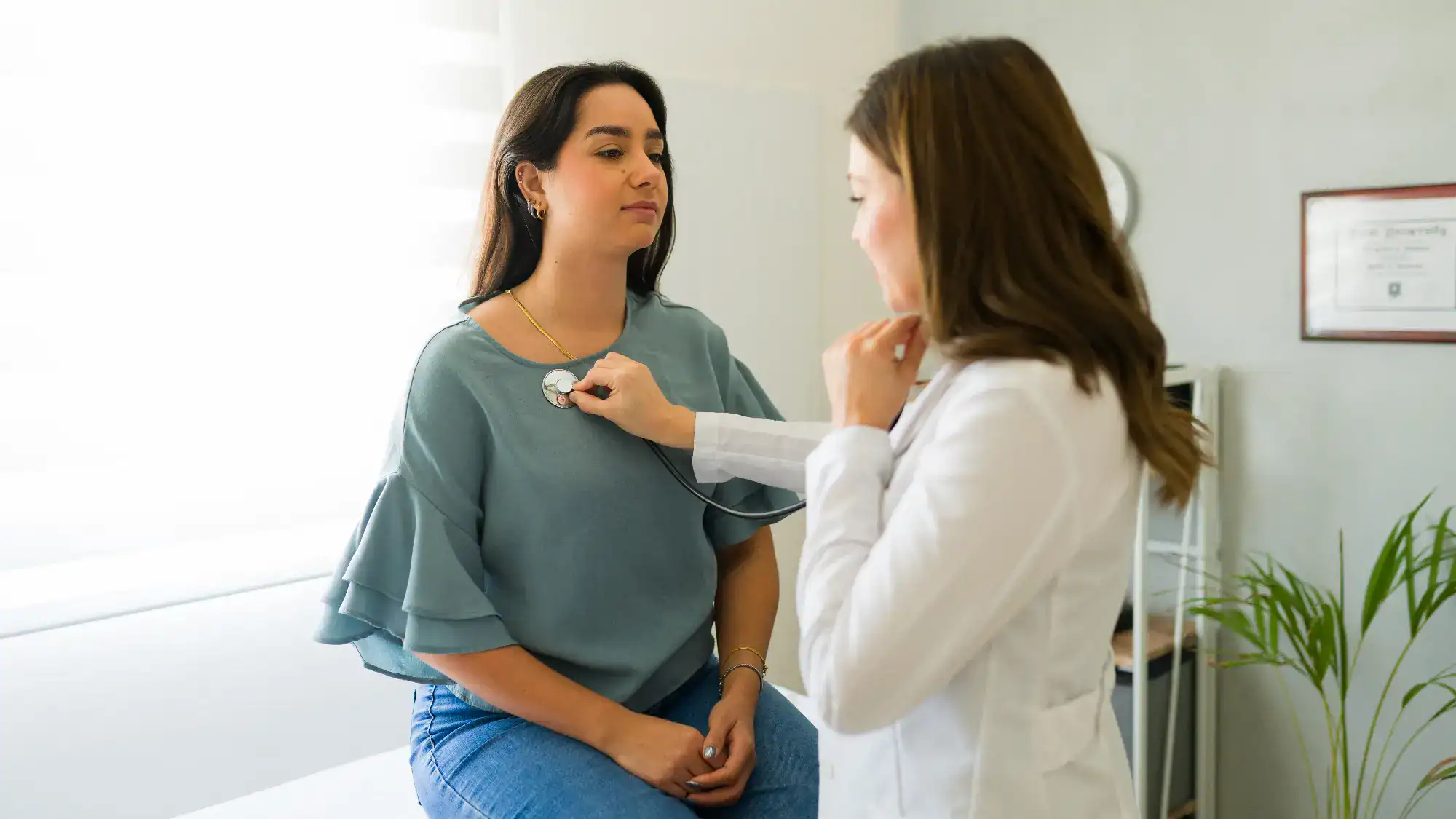 A healthcare professional uses a stethoscope to listen to a woman's chest during a medical examination in a bright, modern FL primary care clinic in Tallahassee. The woman sits calmly on an exam table.