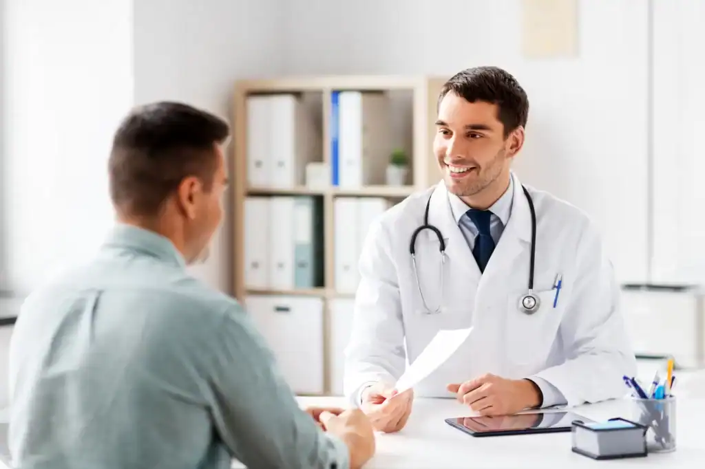 A smiling doctor in a white coat holds a paper and talks with a seated male patient in an office, highlighting quality primary care in Tallahassee, FL. Shelves with binders and medical items are visible in the background.