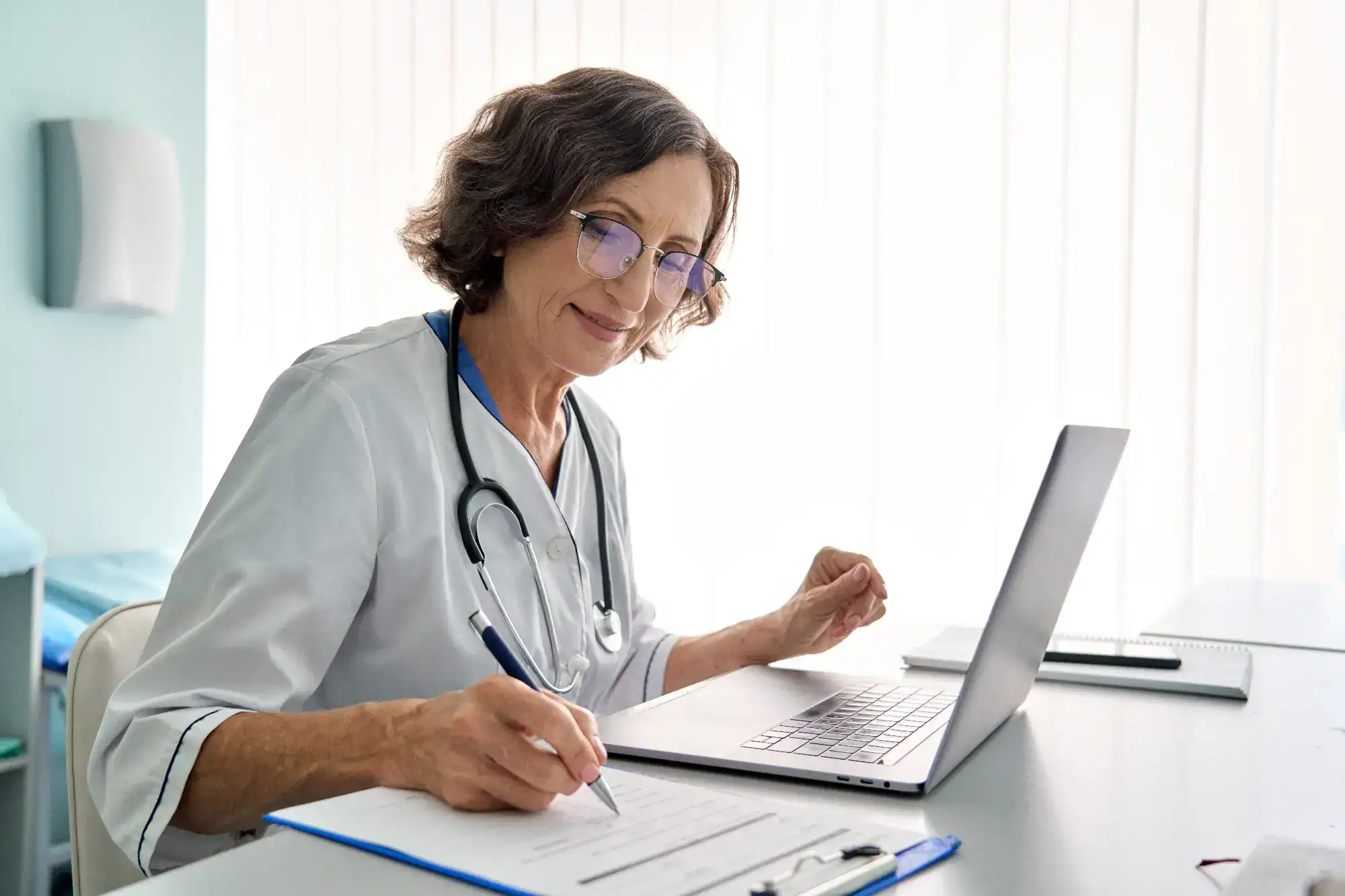Smiling older female doctor with glasses and a stethoscope writes on a clipboard while sitting at a desk with a laptop in a bright FL primary care Tallahassee medical office.