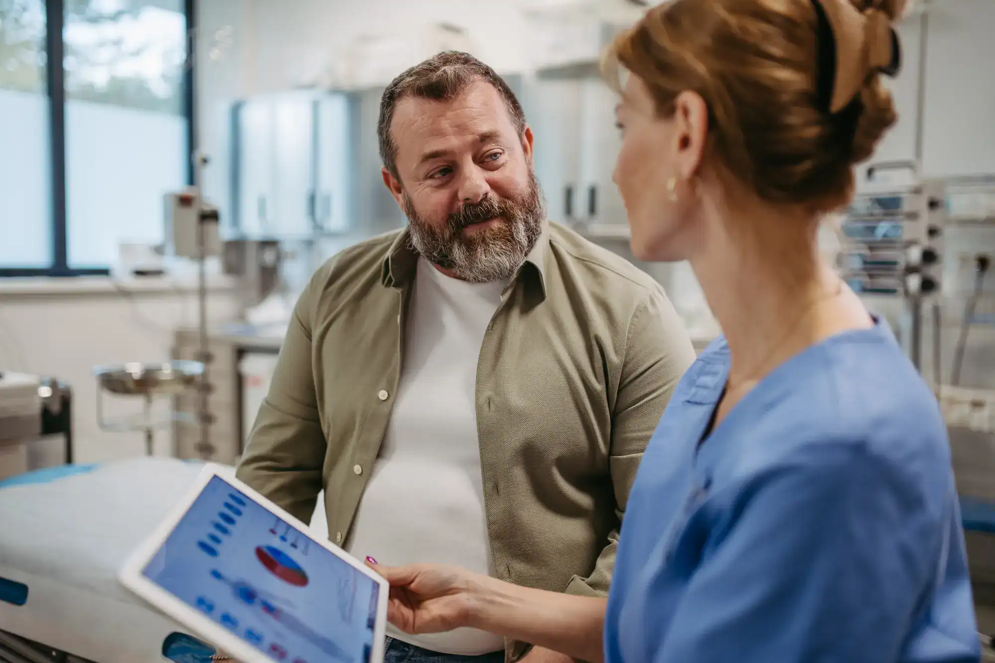 A healthcare worker in blue scrubs shows a tablet with charts to a seated man with a beard in a FL primary care Tallahassee medical office. They appear to be having a focused discussion about the information on the screen.