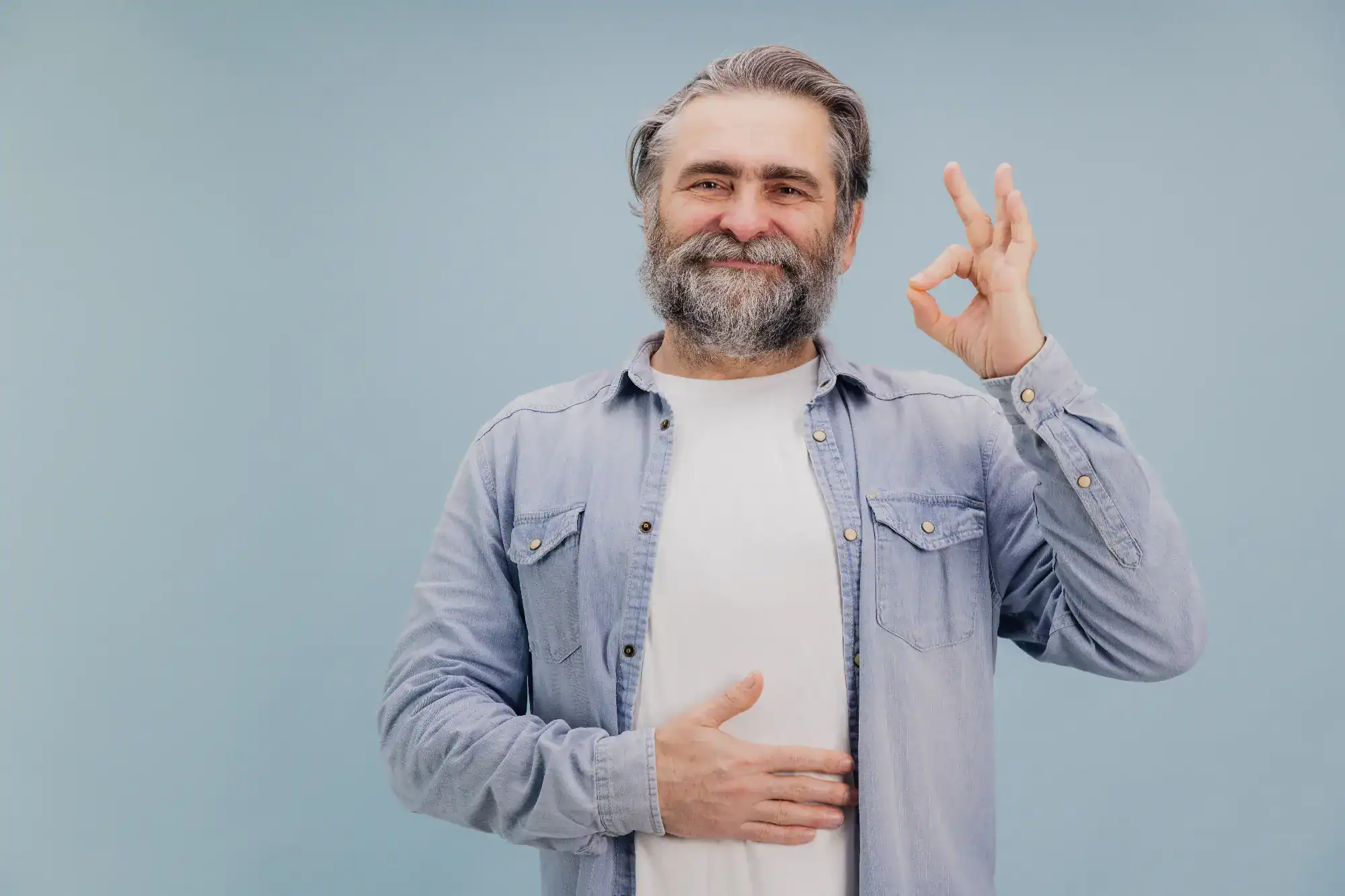 A middle-aged man with a beard, wearing a light blue shirt over a white t-shirt, smiles and makes an "OK" hand gesture while resting his other hand on his stomach, embodying confidence in FL primary care Tallahassee, against a light blue background.