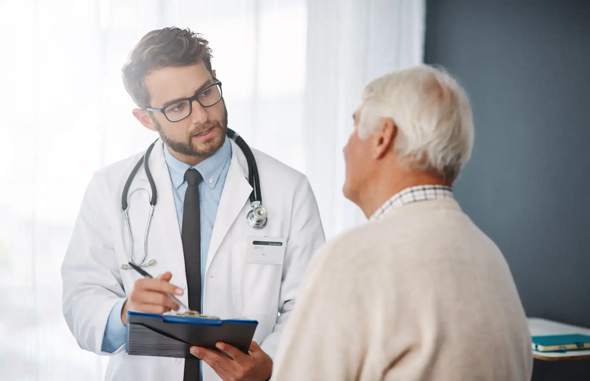 A male doctor with glasses and a stethoscope talks to an older male patient, holding a clipboard and pen while listening attentively in a bright primary care Tallahassee, FL medical office.