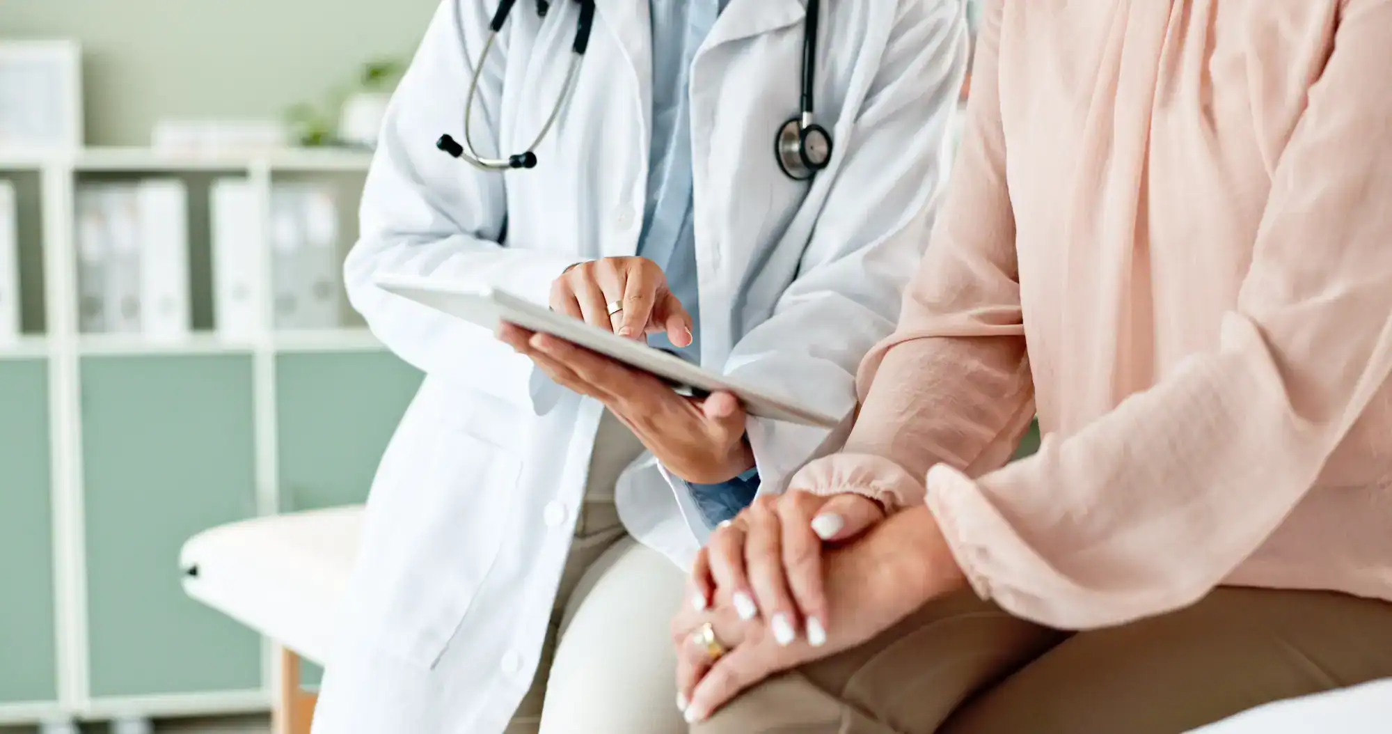 A primary care doctor in Tallahassee, FL, wearing a white coat and holding a tablet, talks to a patient sitting nearby. The patient's hands rest on their lap, with only their torsos and hands visible.
