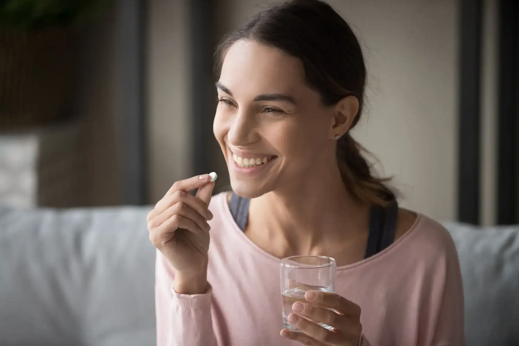 A smiling woman holds a glass of water and a white pill, preparing to take her medication while sitting indoors—reflecting the focus on wellness at FL primary care Tallahassee.