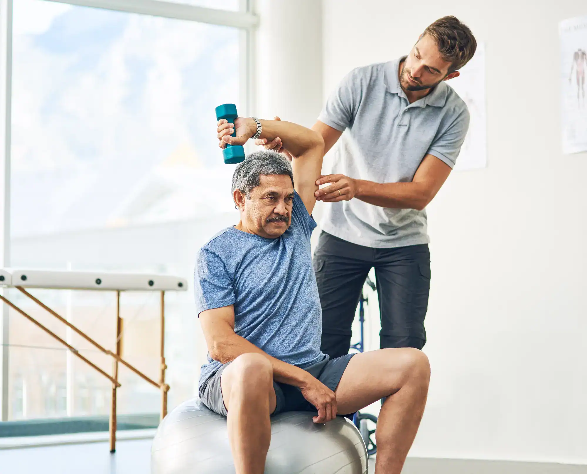 A physical therapist assists an older man, seated on an exercise ball, in lifting a dumbbell overhead during a rehabilitation session at a bright, modern primary care Tallahassee clinic in FL.
