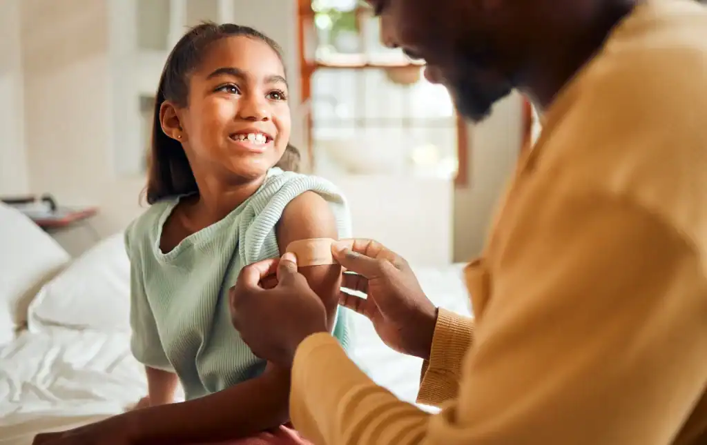 A smiling young girl sits on a bed while an adult gently places a bandage on her upper arm, suggesting she has just received a vaccination or injection at a bright, cozy primary care office in Tallahassee, FL.