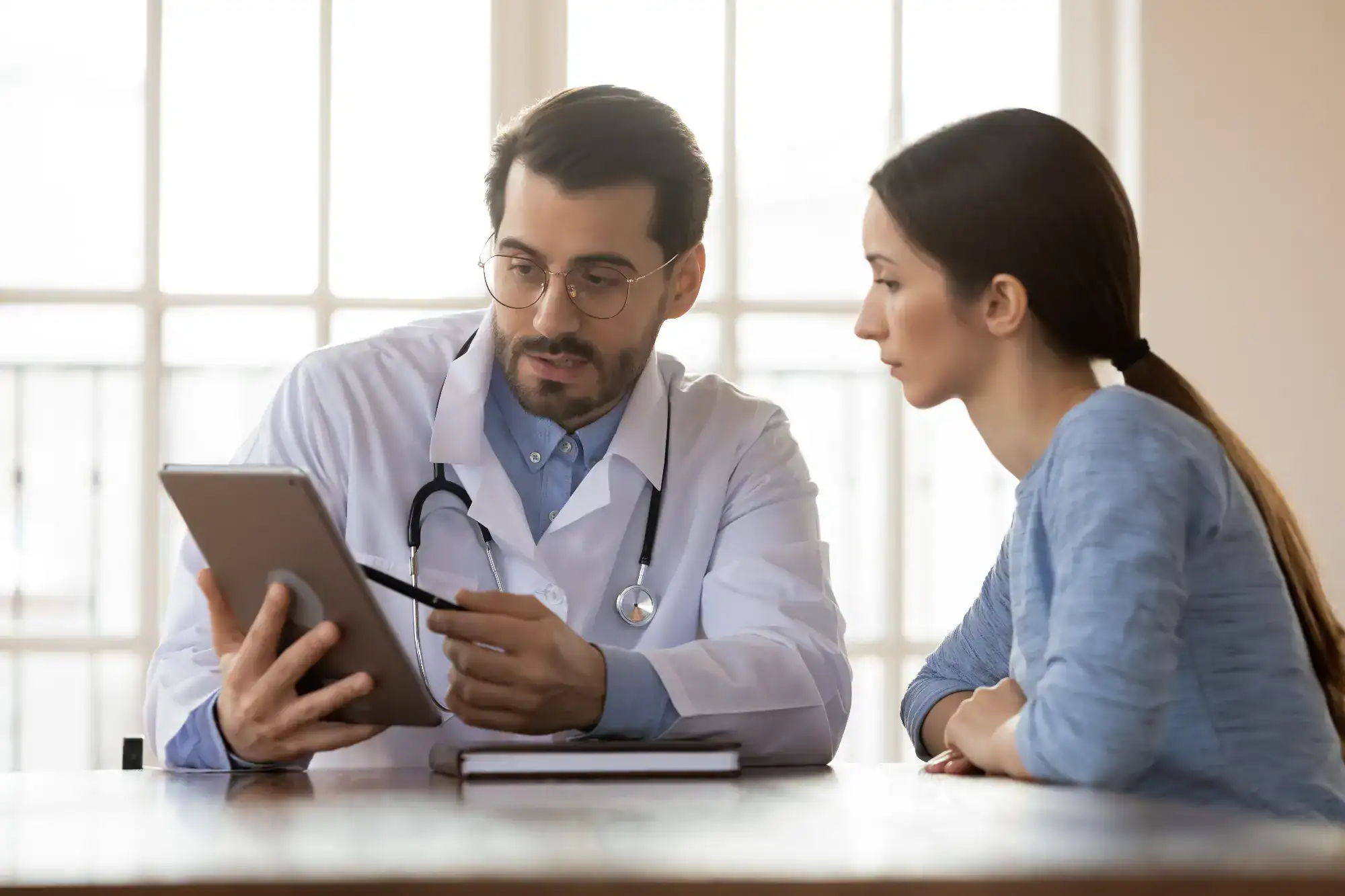 A male doctor in a white coat shows information on a tablet to a woman beside him at a table, discussing medical results in the bright office of a primary care clinic in Tallahassee, FL.