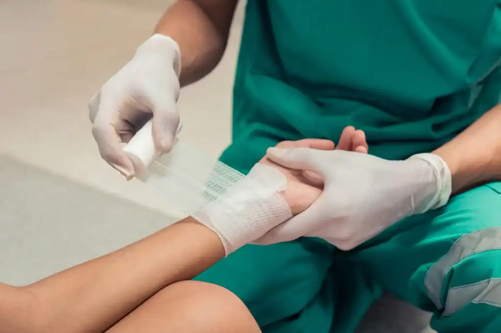 A medical professional in green scrubs and white gloves carefully wraps a patient’s wrist with a bandage, highlighting attentive wound care or first aid treatment in a primary care Tallahassee, FL clinical setting.