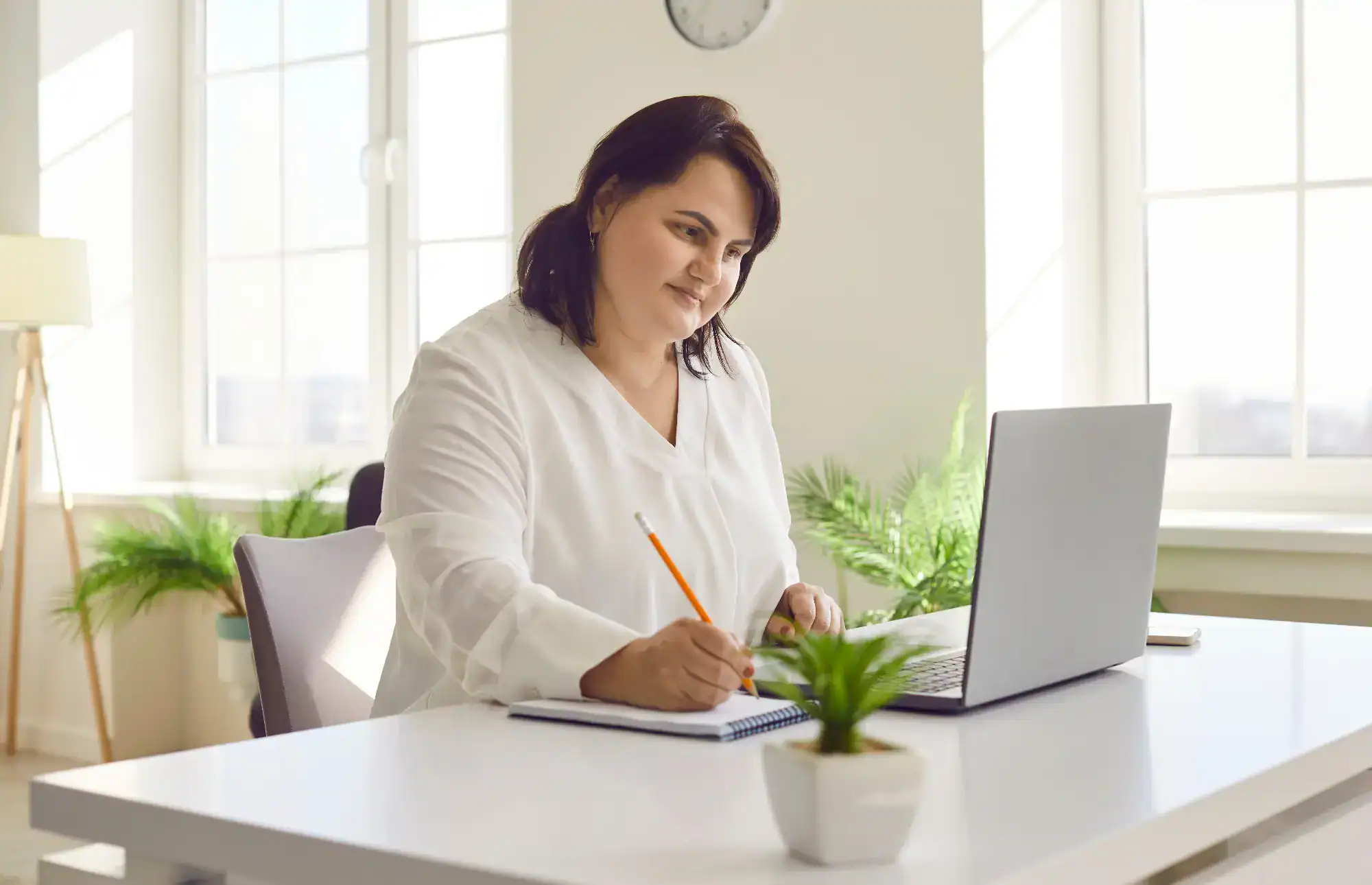 A woman in a white blouse sits at a desk, writing in a notebook and looking at her laptop. The bright room, filled with plants and large windows, sets the scene for primary care work in Tallahassee, FL. A clock hangs on the wall.
