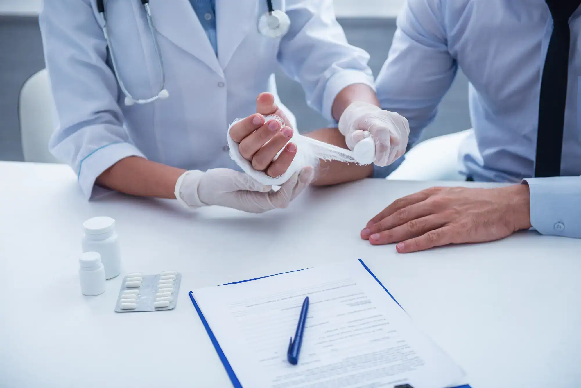 A doctor in a white coat and gloves wraps a patient’s injured wrist with a bandage. Pills, a bottle, and a document with a pen sit on the table—typical care at a primary care Tallahassee clinic in FL.