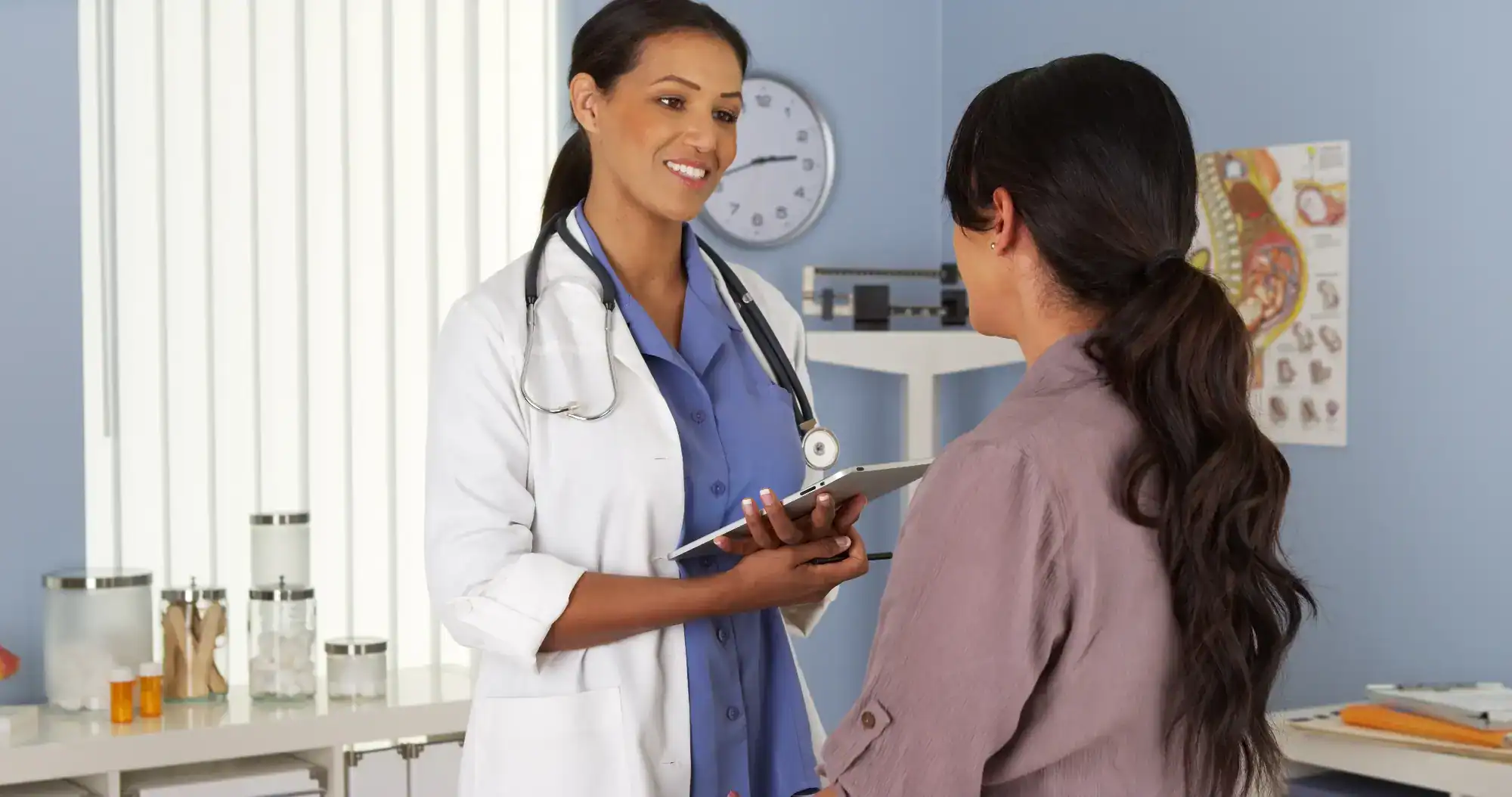 A female doctor in a white coat and stethoscope smiles while talking to a patient in an exam room at a primary care Tallahassee clinic. The doctor holds a tablet, with medical charts and equipment visible in the background.