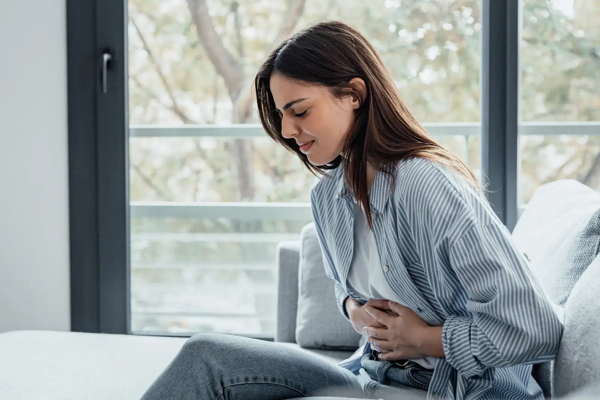 A woman sits on a couch indoors, holding her stomach in discomfort. If you need support, primary care Tallahassee, FL can help address abdominal pain and other health concerns. She wears a striped shirt; trees are visible through the window behind her.