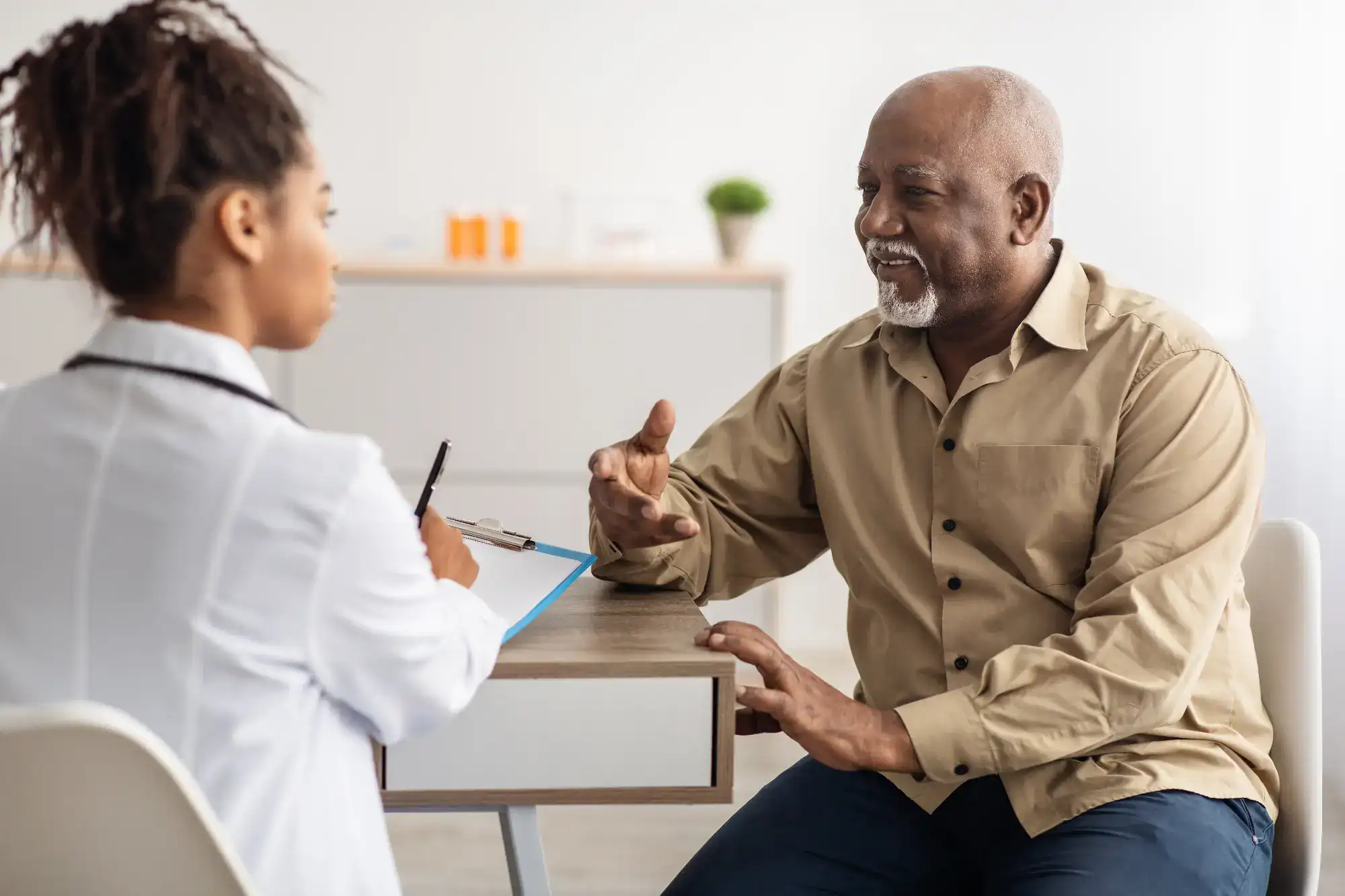 A doctor listens and takes notes while talking to an older man in a beige shirt, who is smiling and gesturing with his hands during a primary care Tallahassee consultation in a bright FL office.