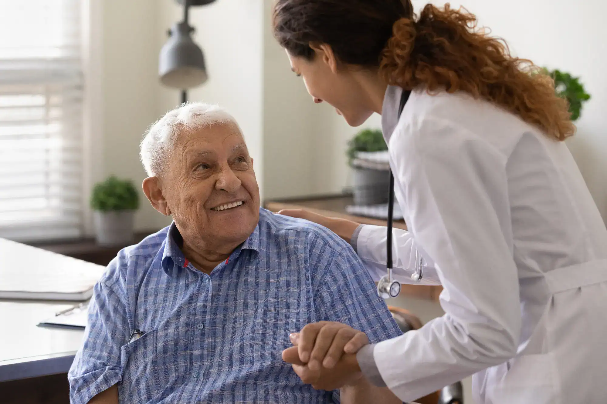 A smiling older man in a blue plaid shirt sits while a female doctor in a white coat holds his hand and speaks with him warmly in a bright, welcoming primary care Tallahassee, FL office.