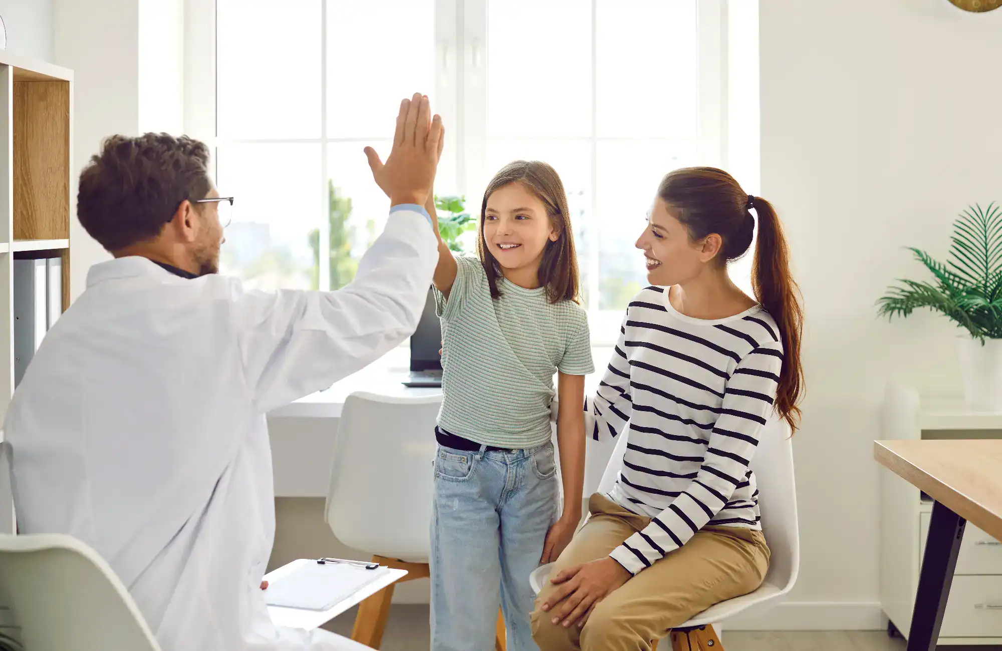 A primary care Tallahassee doctor in a white coat gives a high-five to a smiling young girl, who stands next to a woman sitting on a chair in a bright, modern FL office with large windows and plants in the background.