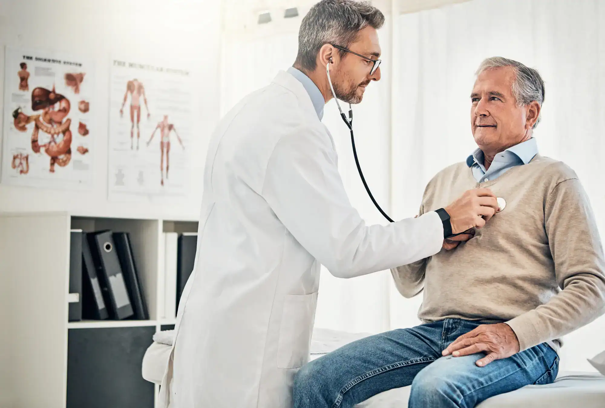 A doctor uses a stethoscope to check the heart of an older man sitting on an exam table in a FL primary care Tallahassee medical office. Medical posters and shelves with folders are visible in the background.