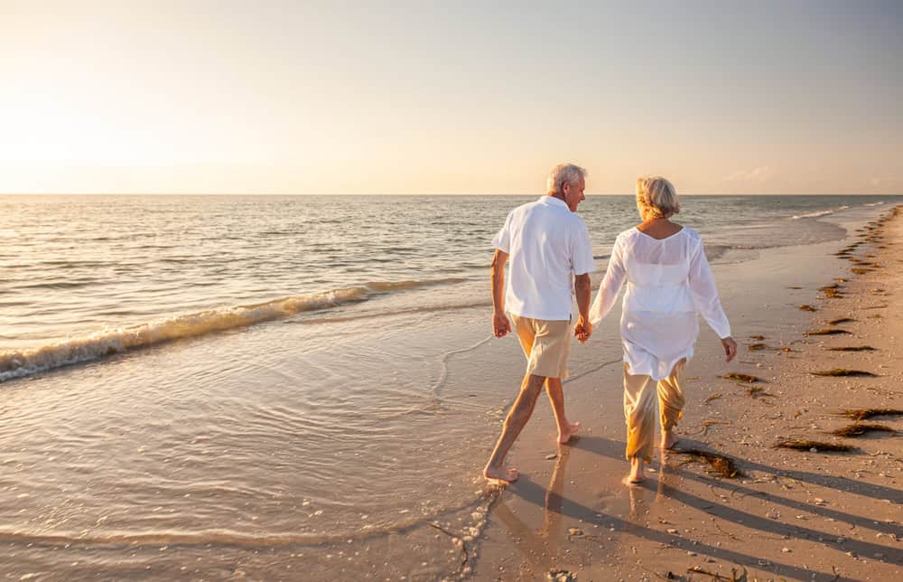 An elderly couple, dressed in light-colored clothes, walks hand in hand along the shoreline of a sandy FL beach at sunset, with gentle waves and seaweed scattered on the sand.