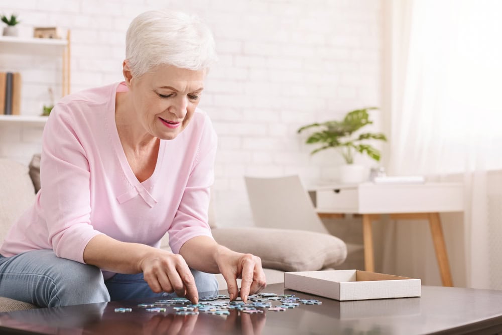 Older woman with short white hair and a light pink sweater sits at a table, smiling while working on a jigsaw puzzle in her bright, cozy Tallahassee, FL living room; she enjoys staying sharp and healthy with quality primary care.