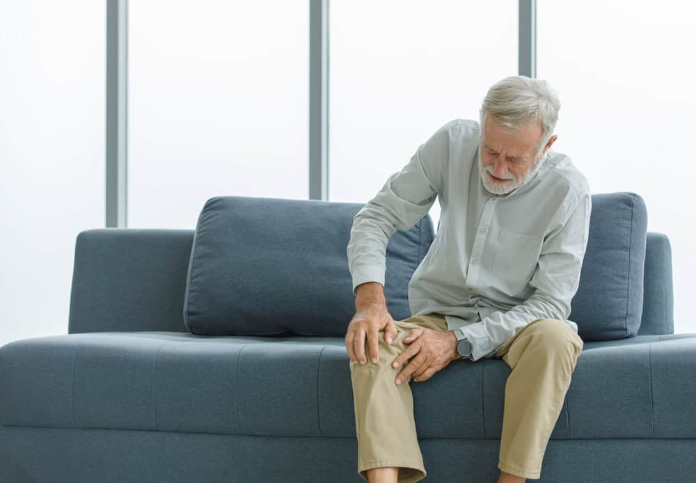 An older man sits on a blue sofa in FL, wearing a light shirt and beige pants, holding his knee with a pained expression, suggesting discomfort or pain that may require primary care Tallahassee attention.