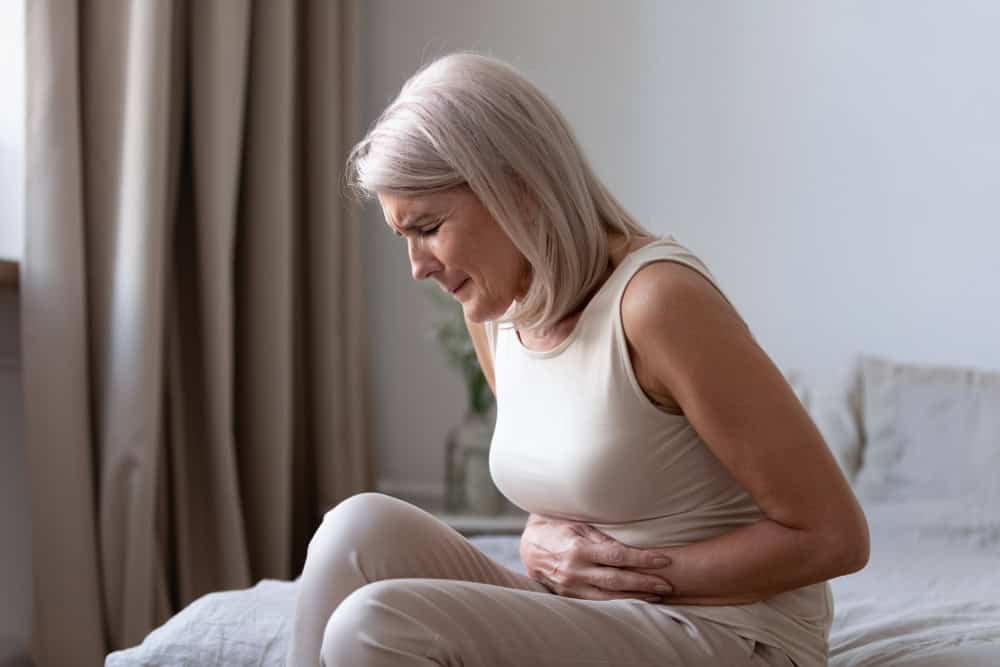 An older woman with gray hair sits on a bed in a softly lit room, clutching her stomach and wincing in pain. If you’re experiencing discomfort like this, consider reaching out to primary care Tallahassee, FL for support and treatment.