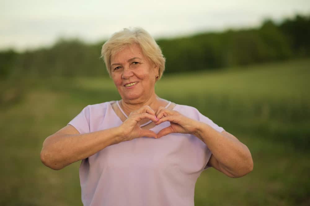 Older woman with short blonde hair, wearing a light purple shirt, stands in a green FL field near Tallahassee, smiling and making a heart shape with her hands—celebrating well-being and primary care.