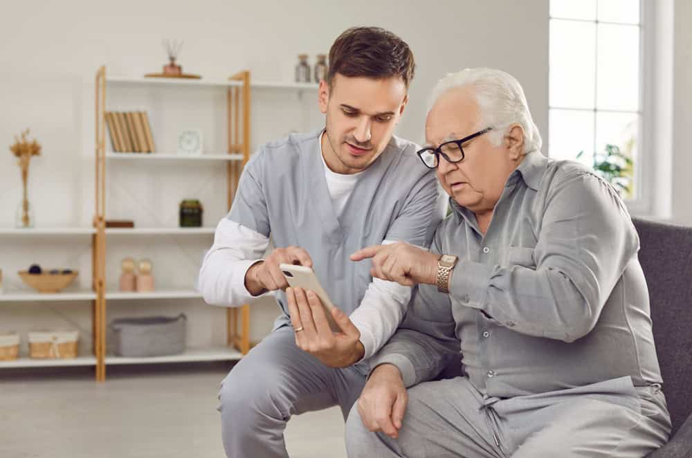 A young male caregiver in scrubs helps an elderly man with white hair and glasses use a smartphone as they sit together on a couch in a bright, homey living room, reflecting the warmth of FL primary care Tallahassee.