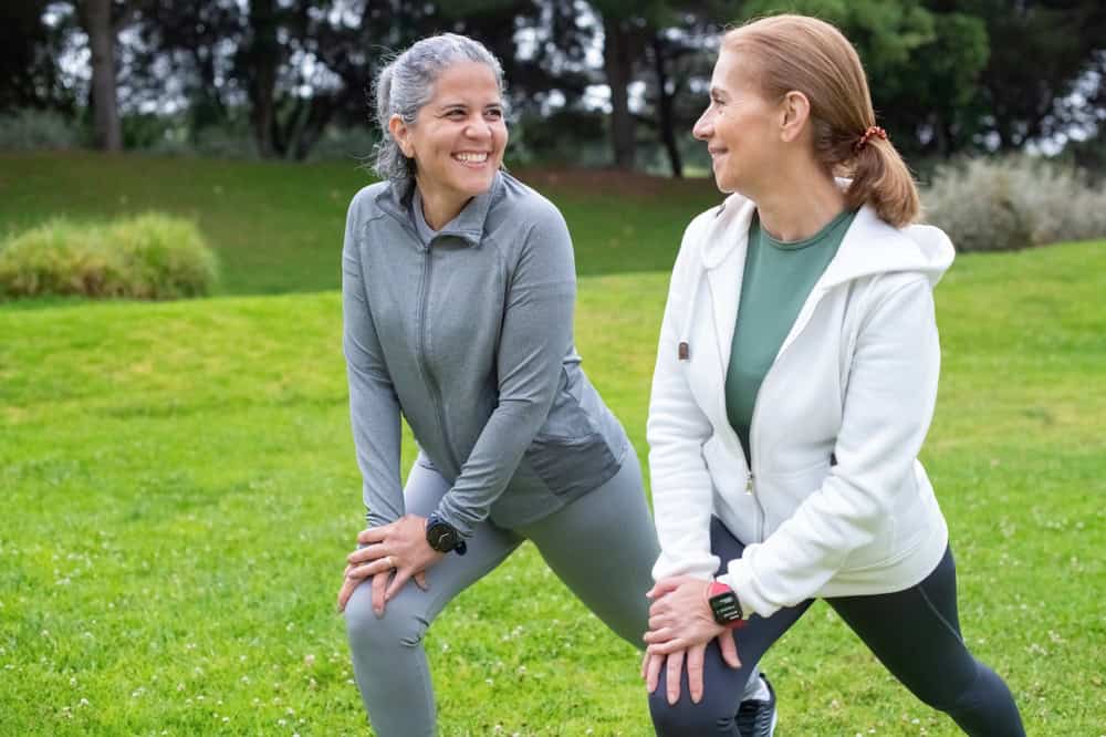 Two women in athletic wear smile at each other while stretching outdoors on a grassy field with trees in the background—enjoying healthy living inspired by primary care in Tallahassee, FL.