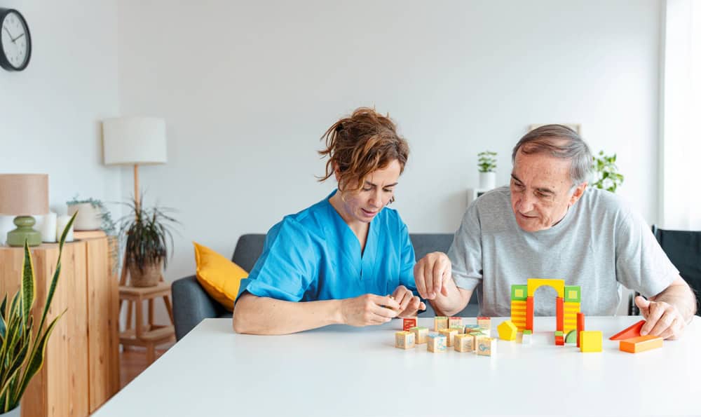 A caregiver in blue scrubs and an older man sit at a table building a colorful structure with wooden blocks in a bright, cozy living room, illustrating the personalized support found in primary care Tallahassee, FL.